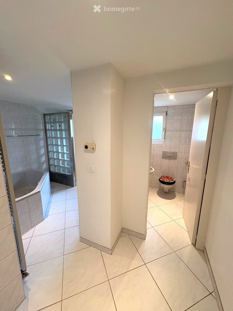 Hallway with tiled floors and white walls, leading to a bathroom with a toilet and a bathtub.