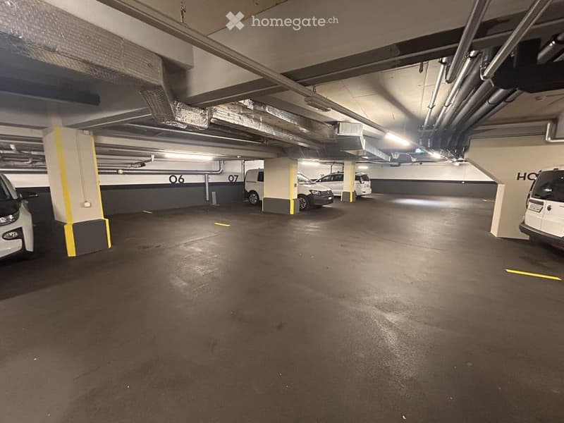 Underground parking area with several parked cars, marked parking spaces, and structural pillars. The area is well-lit with fluorescent lighting and has a concrete floor.