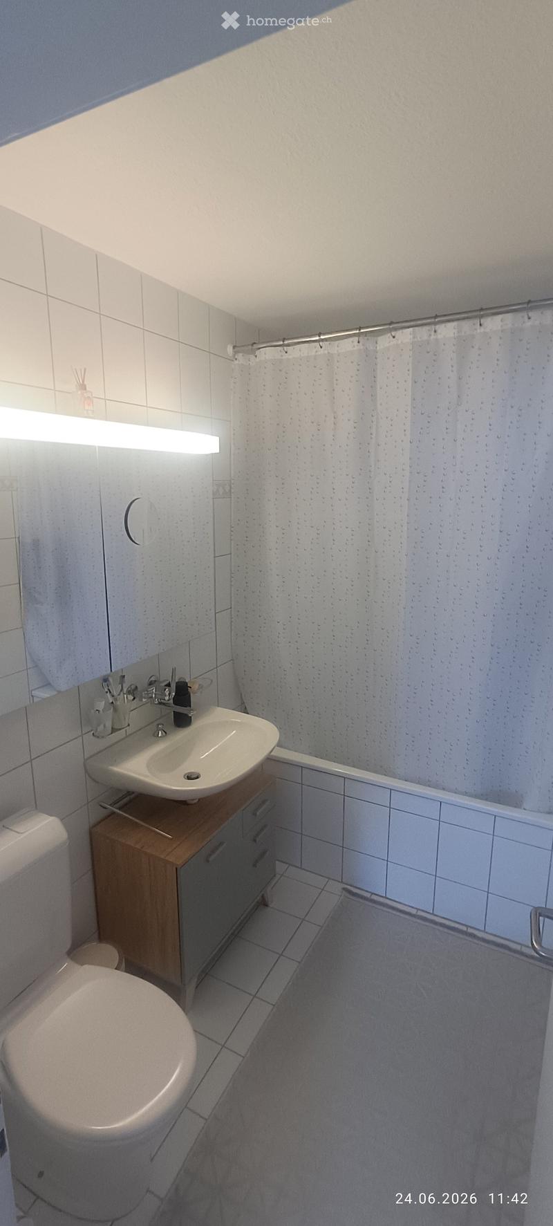 Modern bathroom with white tiles, a toilet, a sink with a wooden cabinet, a mirror, and a shower area with a curtain.