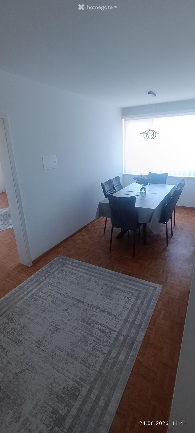 Dining room with wooden flooring, a large dining table surrounded by six chairs, a table runner, and a vase with flowers on the table. A patterned rug is placed on the floor near the table.