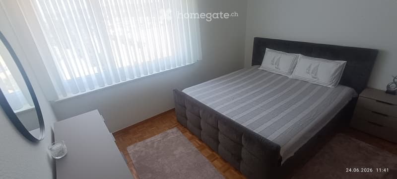 Modern bedroom with a grey upholstered bed, matching pillows, a nightstand with a decorative item, a small table with a candle, and a grey rug on a wooden floor.
