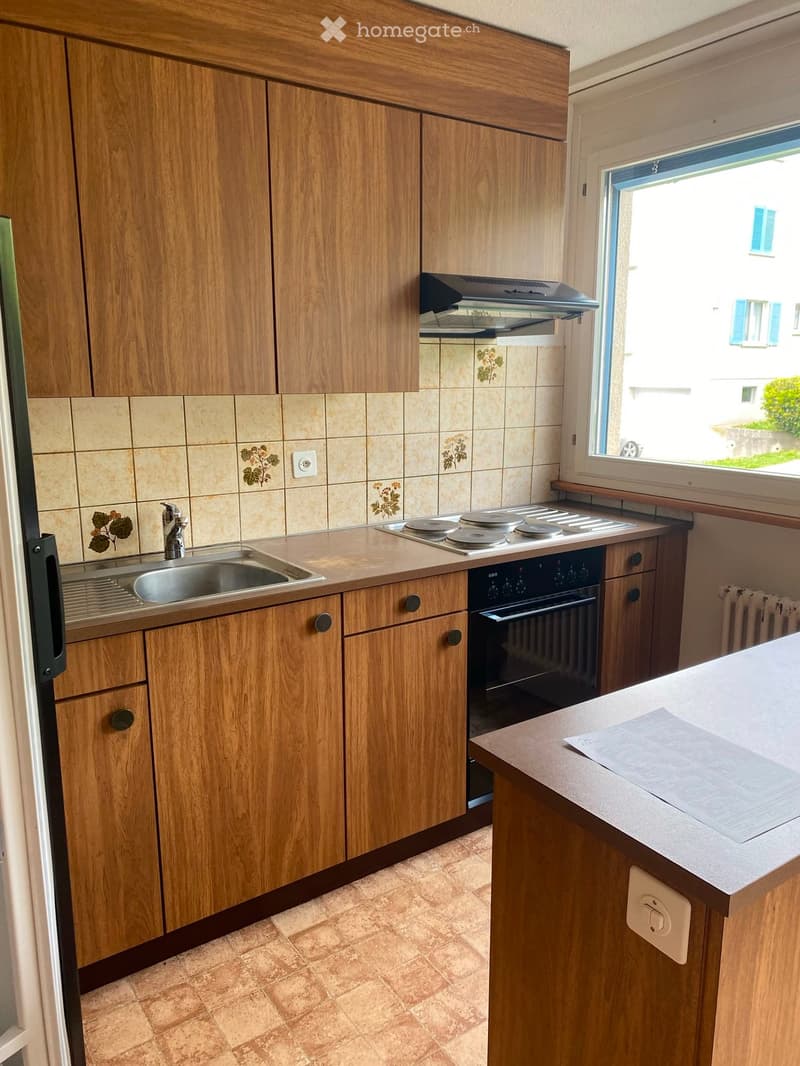 A modern kitchen with wooden cabinets, a stainless steel sink, a stove, and an oven. The countertops are light-colored, and the backsplash features floral tiles.