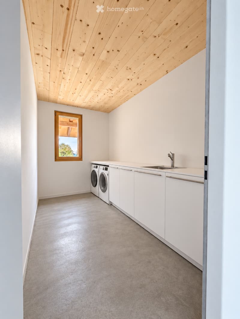 Modern, minimalist laundry room with white cabinets, a sink, and a washing machine. Features a skylight window and a wooden slanted ceiling.