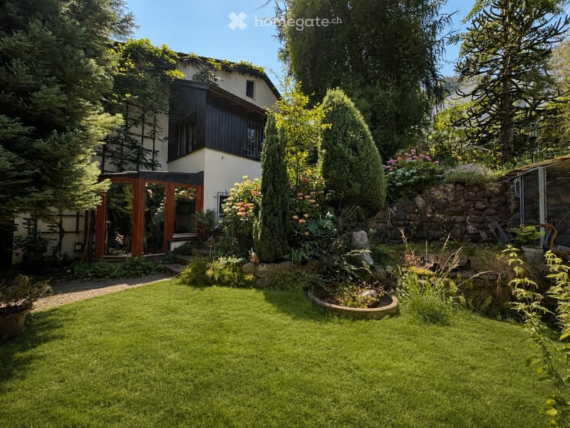 Well-maintained garden with a lawn, various plants, trees, and a stone wall. The house in the background has a modern design with large windows and a wooden door.