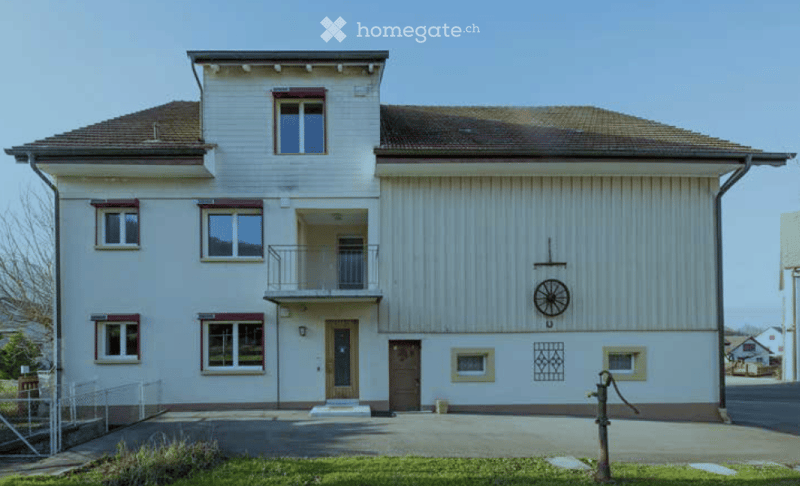 Two-story building with white siding, multiple windows, a small balcony, and a decorative wheel on the wall.