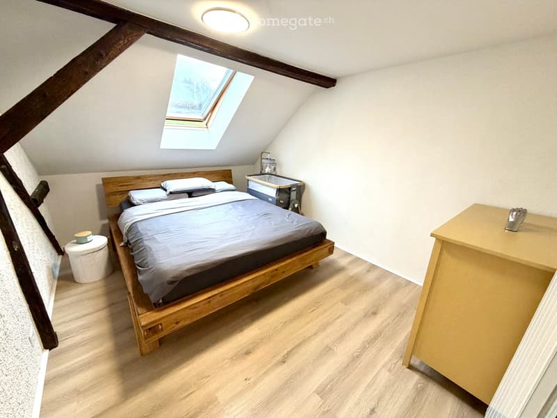 Cozy attic bedroom with wooden bed, nightstand, small stool, baby crib, and a small table with a decorative item.
