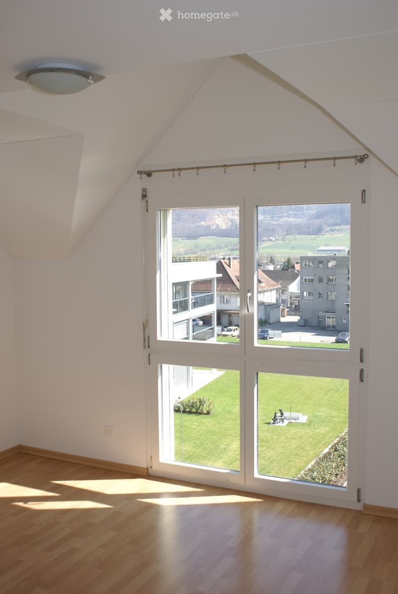 Bright, empty room with wooden flooring and large windows offering a scenic view of a green landscape and buildings. The room features white walls and a modern ceiling light fixture.