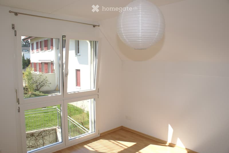 Bright, empty room with large windows and sliding doors leading to a balcony and garden view. Light wooden flooring and white walls.