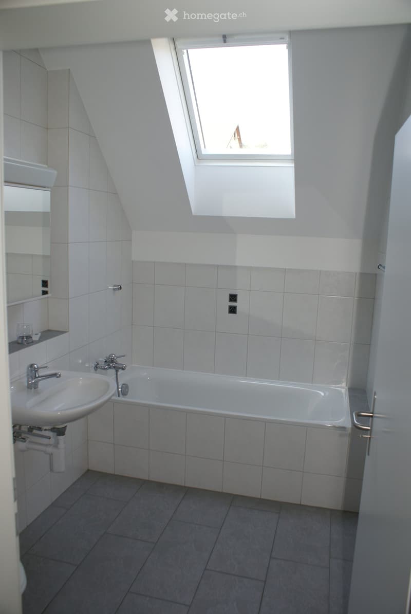 Modern bathroom with a bathtub, sink, and mirror. The room features white tiled walls and a skylight window providing natural light.