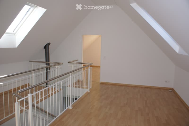 Bright, empty hallway with wooden flooring, white walls, and a modern staircase featuring a white metal railing and a black central column. Skylights provide natural light, and a doorway leads to another room.