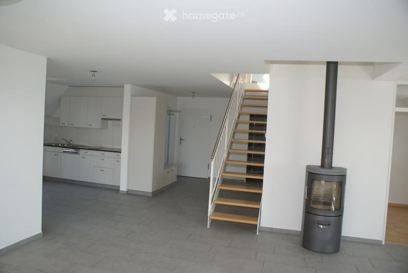 Open-plan living space with a staircase leading to an upper level, a modern kitchen area with white cabinets and stainless steel appliances, and a wood stove with a chimney. The floor is tiled in the main area and wooden in the adjacent section.