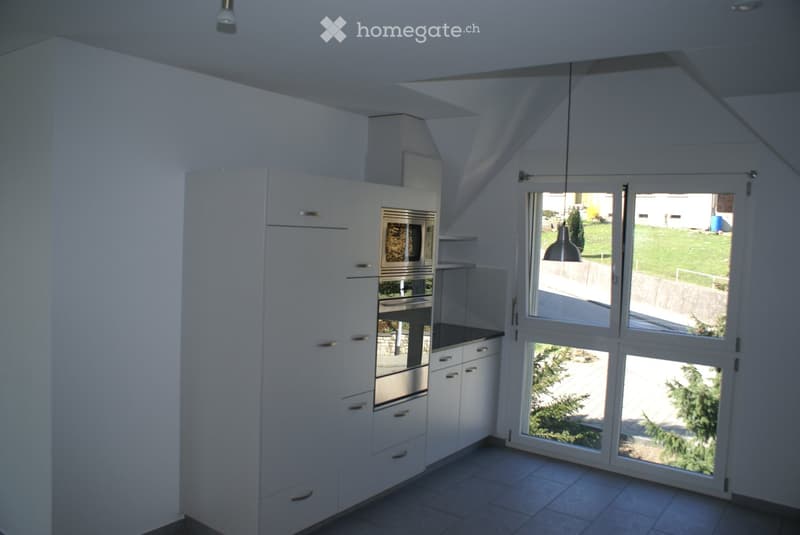 Modern kitchen with white cabinets, stainless steel oven, and large windows overlooking a green area.