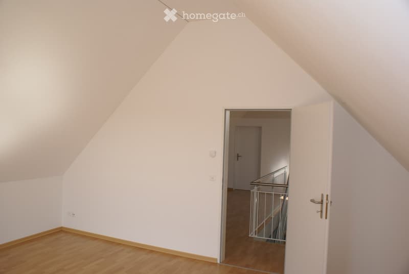 An empty attic room with wooden flooring, white walls, and a slanted ceiling. An open doorway leads to a hallway with a staircase visible in the background.