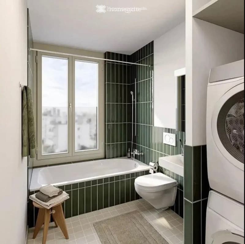 Modern bathroom with a bathtub, toilet, sink, and washing machine. Green tiled walls, large windows, and a small wooden stool with towels.