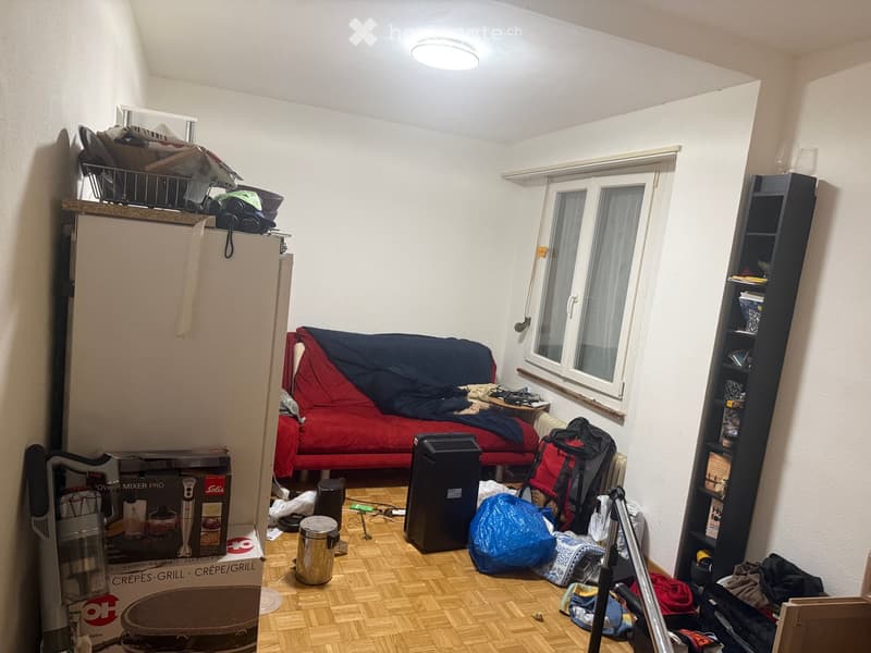 A small, furnished room with a sofa, refrigerator, shelf, tripod, backpack, and various items scattered around.