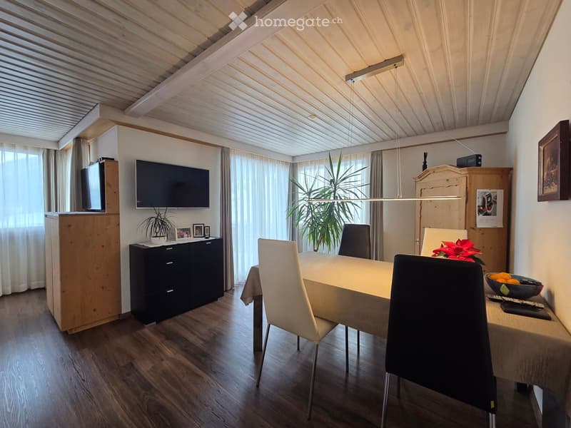 Dining room with a wooden dining table, white chairs, a television mounted on the wall, a cabinet, and a plant in the corner.