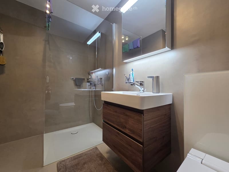 Modern bathroom with a white sink, wooden vanity, large mirror, and glass-enclosed shower. Includes a toothbrush holder and a towel on the floor.