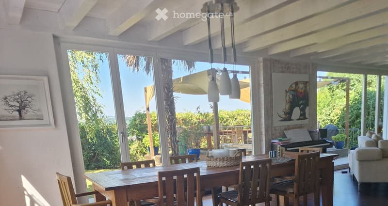 Bright, airy room with large windows and a view of greenery. Features a wooden dining table with chairs, a sofa, a piano, and decorative items including a framed picture and a basket on the table.