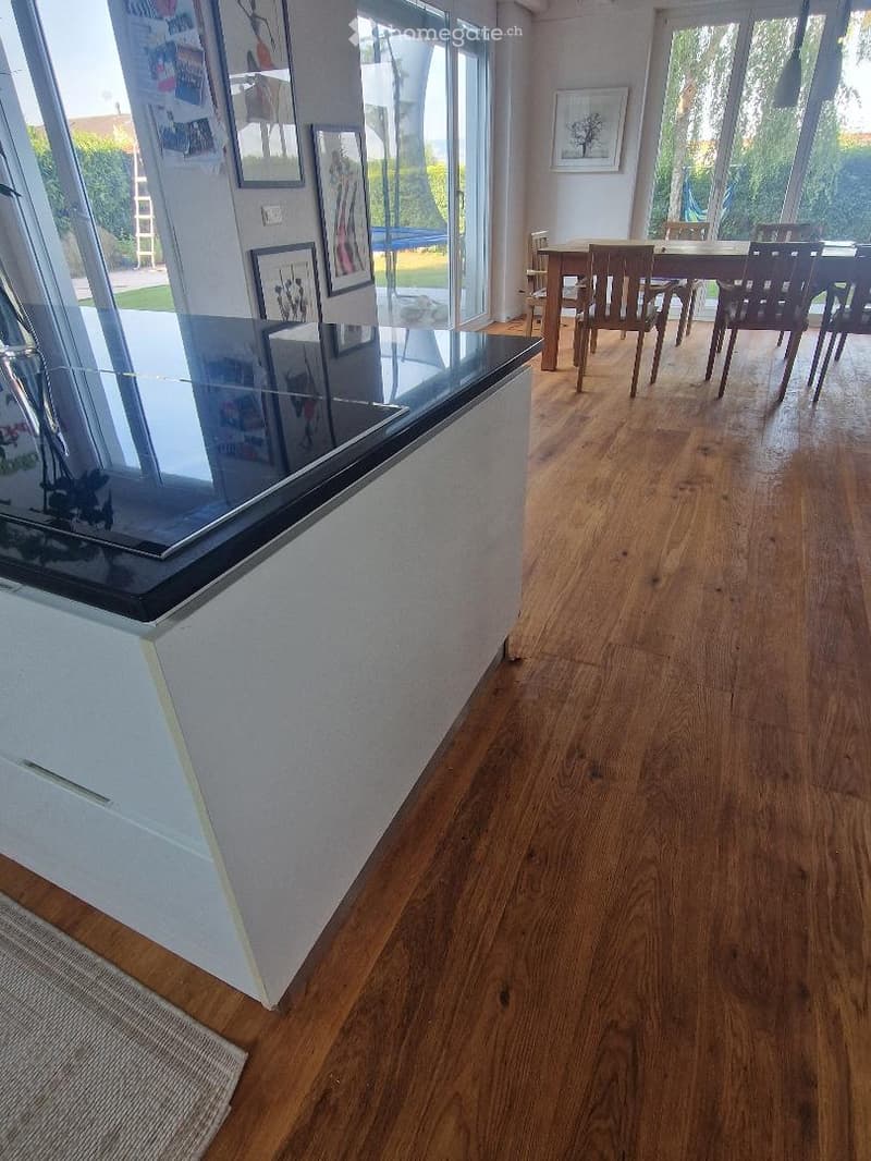 Open-plan living and dining area with wooden flooring, large windows, and a dining table with chairs.