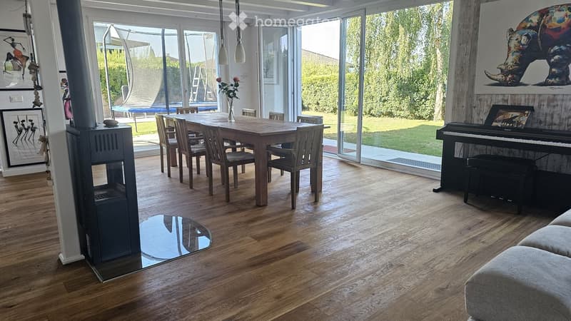 Bright, open-plan living and dining area with wooden flooring, large windows, a dining table with chairs, a piano, and artwork on the walls.