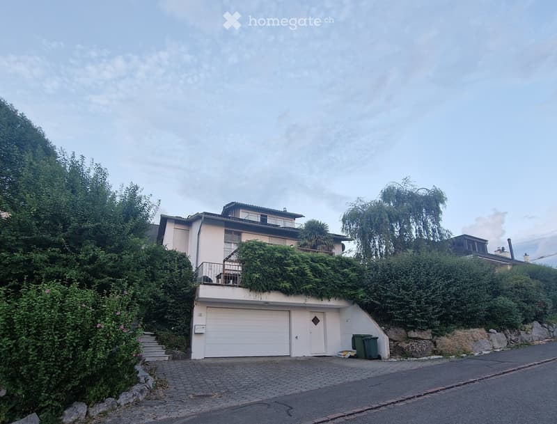 Two-story house with a garage, balcony, and lush greenery on the roof and surrounding area.