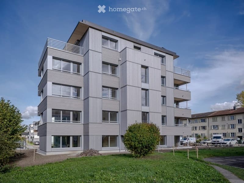 Modern multi-story apartment building with grey facade, large windows, and balconies. Surrounded by green lawn and parking area.