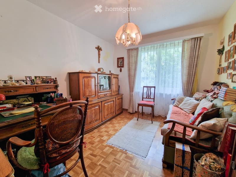 A cozy living room with wooden flooring, a sofa with pillows, a wooden cabinet, a chair, a small table, and a hanging lamp.