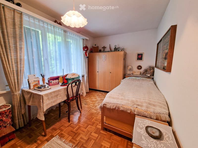 Cozy bedroom with wooden flooring, a single bed with a white bedspread, a wooden table with a chair, a wooden cabinet, and a lamp on a nightstand.