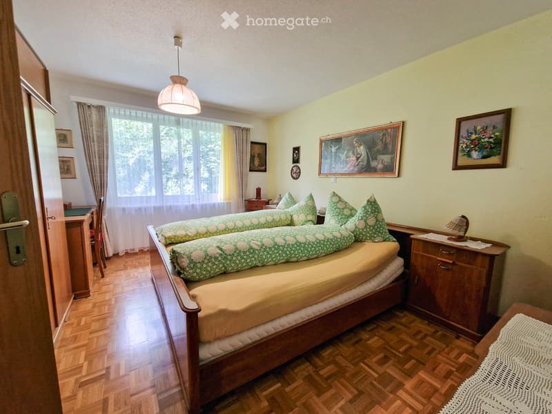 Cozy bedroom with wooden flooring, a large bed with green and white patterned pillows, a nightstand with a lamp, a small table with a chair, and several framed pictures on the walls.