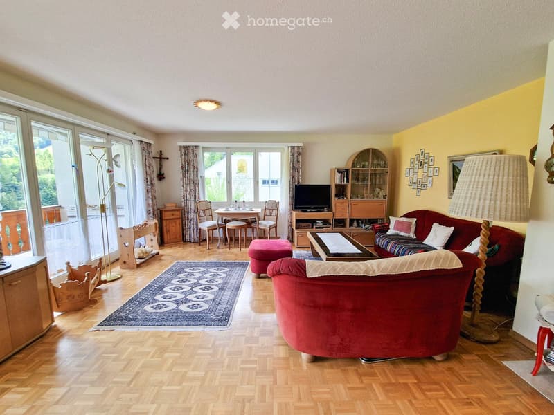 Bright living room with wooden flooring, large windows, and a view of greenery. Features a red sofa, coffee table, ottoman, dining area with chairs and table, TV cabinet, and decorative items.