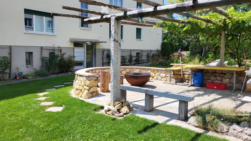 Stone wall, wooden bench, fire pit, wooden table, blue barrel, red crate, green grass, stone path