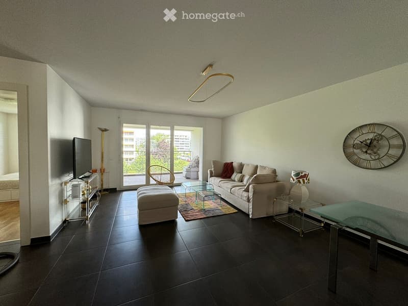 Modern living room with dark tiled flooring, white walls, a large sofa with cushions, an ottoman, a glass coffee table, a wicker chair, a television on a stand, a floor lamp, and a decorative wall clock. Large sliding glass doors lead to a balcony.