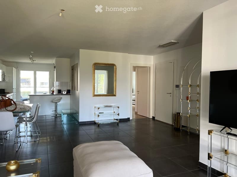 Modern living room with a sofa, coffee table, chairs, a stool, a mirror, and a television. The room features a sleek design with dark tiled flooring and white walls.