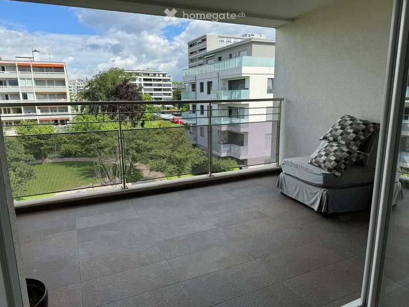 Balcony with tiled floor, modern metal railing, grey sofa with black and white checkered pillows, overlooking a cityscape with modern buildings and greenery.