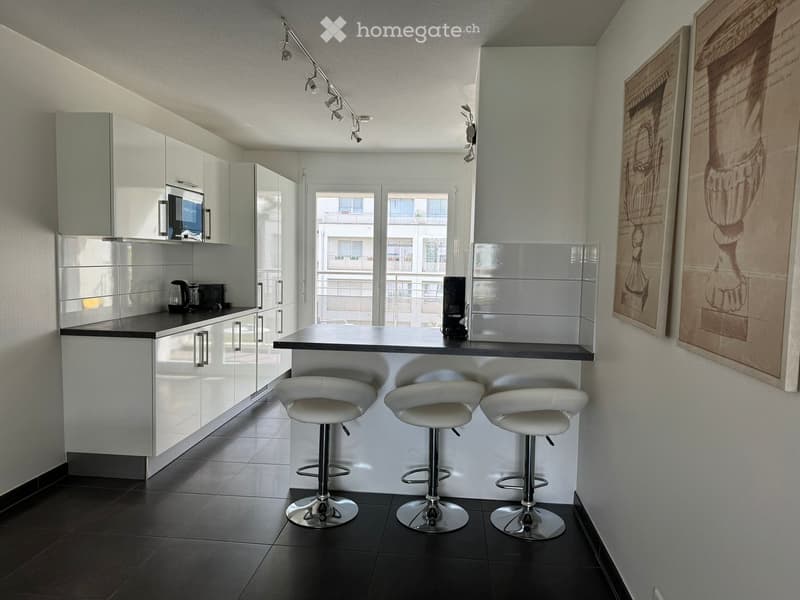 Modern kitchen with white cabinets, black countertops, and three white bar stools at a central island. Features a microwave, coffee maker, and a refrigerator, with framed artwork on the wall.