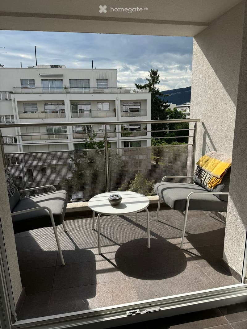 Balcony with a view of a modern building and mountains, featuring a round table, two chairs, and a sofa with a blanket.