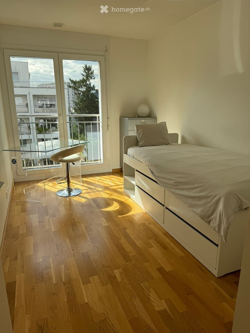 Modern bedroom with wooden flooring, a bed with a grey pillow, a glass table, and a white chair near a large window with a balcony view.
