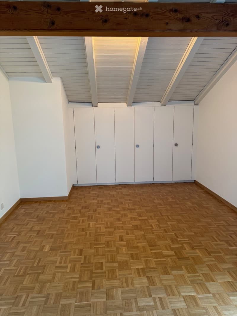Empty room with wooden herringbone flooring, white walls, and built-in white cabinets. The ceiling features exposed wooden beams and recessed lighting.