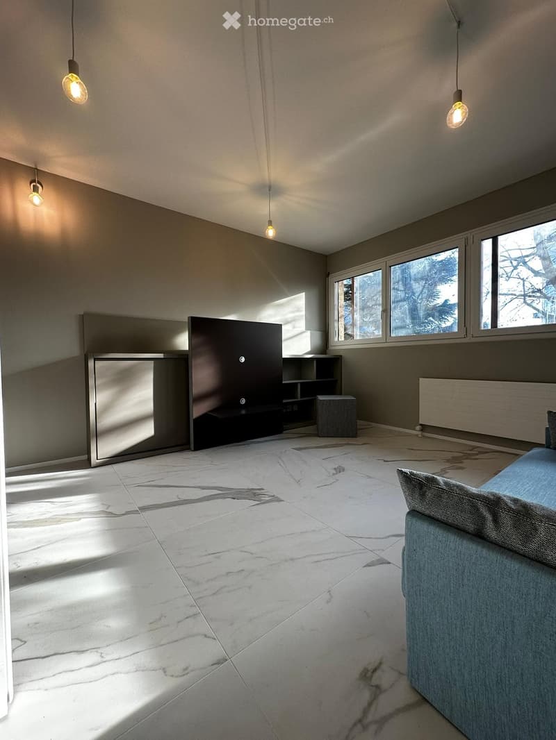 Modern living room with marble flooring, large windows, a grey sofa, a small stool, a built-in shelf unit, and a sleek fireplace.