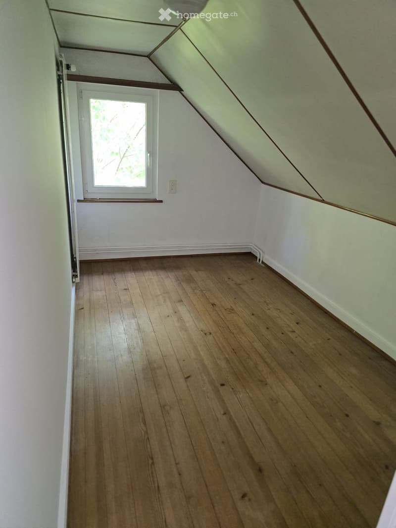 Empty attic room with wooden flooring, white walls, and a slanted ceiling. A small window with a view of greenery is visible, along with a light switch and a door frame.