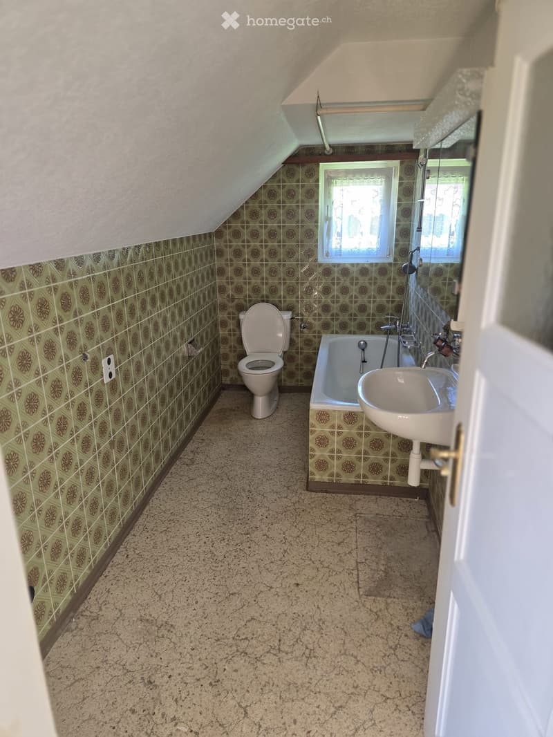 Bathroom with green patterned tiles, a toilet, a bathtub, and a sink. The room has a slanted ceiling and a window letting in natural light.