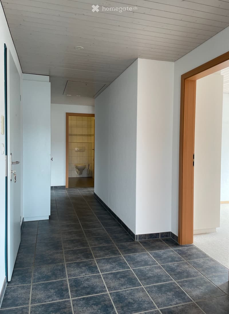A narrow hallway with dark tiled flooring and white walls, featuring a partially open door leading to a small bathroom with a toilet and tiled walls.