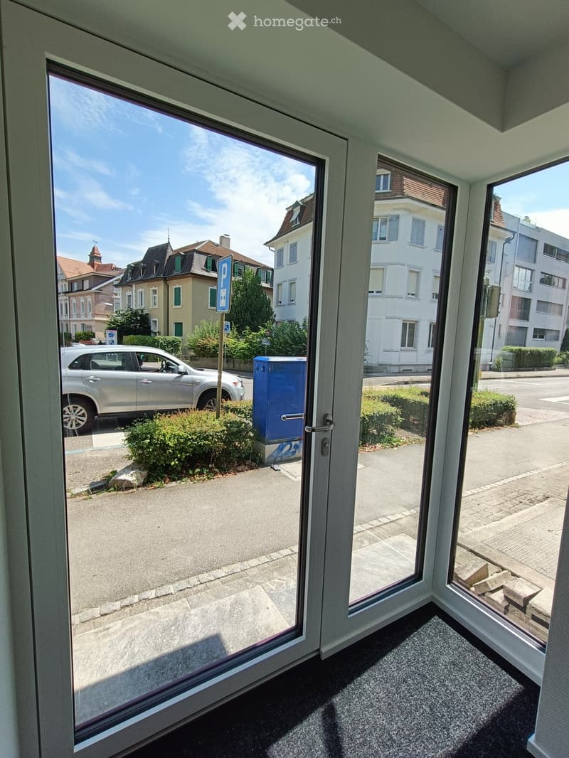View from inside a room through large glass doors showing a street with parked cars, buildings, and a parking sign.