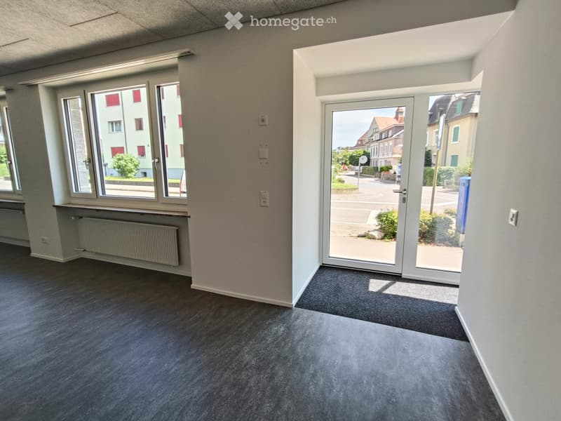 Empty room with grey carpeted floor, white walls, large windows, and a sliding glass door leading to a view of the street.