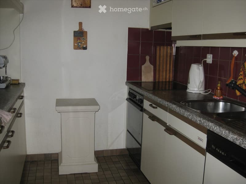 A compact kitchen with white cabinets, granite countertops, and a tiled backsplash. Features include an oven, sink, electric kettle, and a coffee machine.