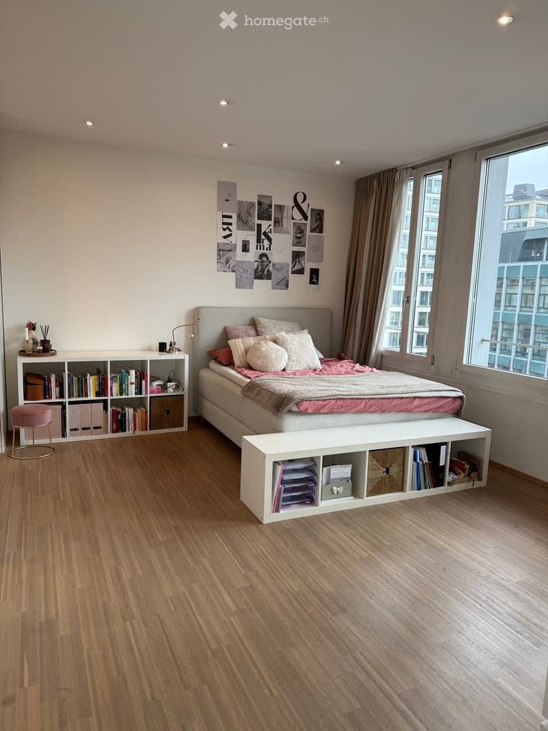 Modern bedroom with wooden flooring, large window with city view, grey bed with pink bedding, white bench, bookshelf, and decorative items.