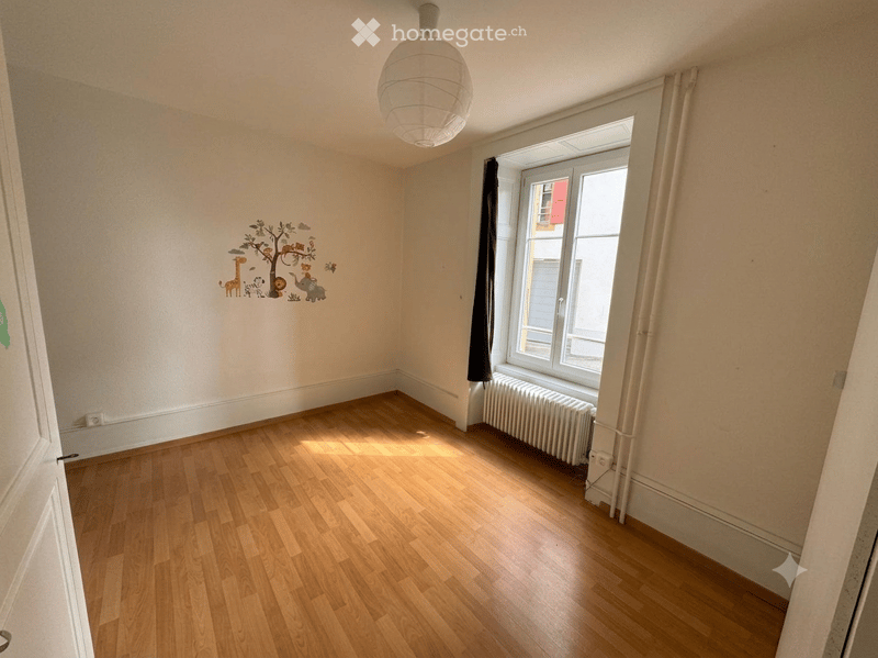 Empty room with wooden flooring, white walls, a window with curtains, a radiator, and a decorative mural of animals and a tree on one wall.
