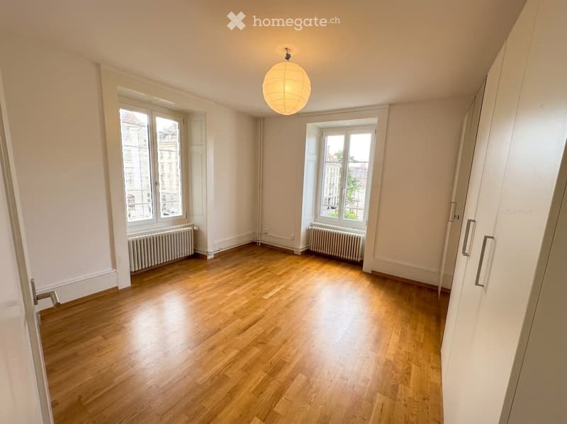 Empty room with wooden flooring, white walls, large windows, radiators, and a hanging light fixture.