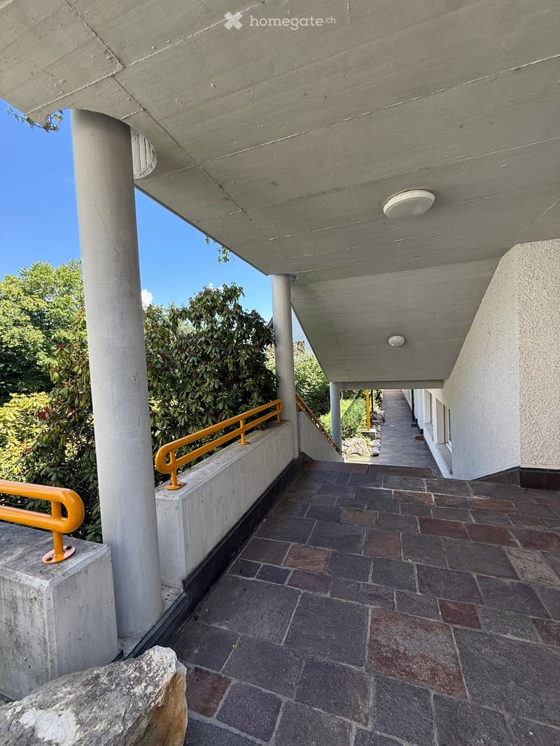 A covered outdoor corridor with a tiled floor, concrete columns, and yellow handrails, leading to a garden area.
