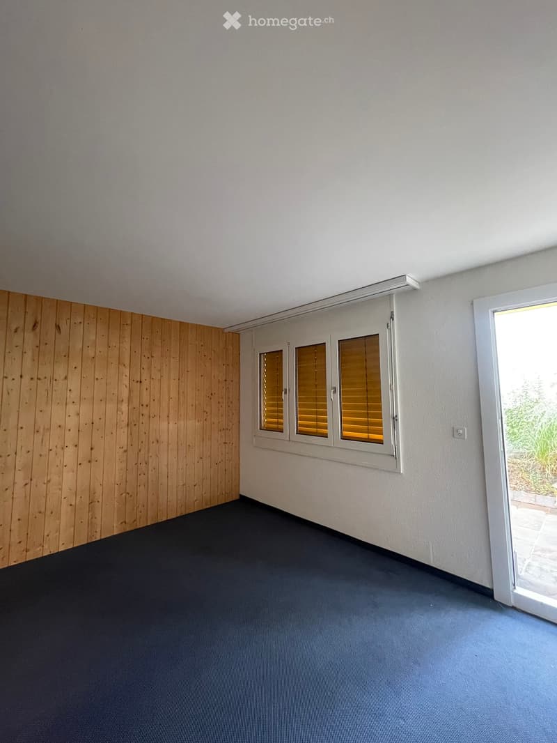 Empty room with wooden panel wall, carpeted floor, large window with blinds, and an open door leading outside.